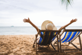 Pessoa com chapéu de palha relaxando em uma cadeira de praia colorida, de frente para o mar, com os braços abertos. Ela está cercada por areia, água tranquila e um coqueiro ao lado.