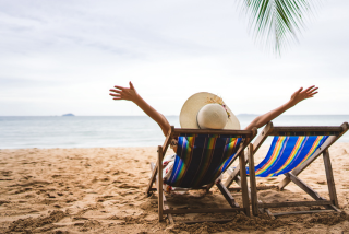 Pessoa com chapéu de palha relaxando em uma cadeira de praia colorida, de frente para o mar, com os braços abertos. Ela está cercada por areia, água tranquila e um coqueiro ao lado.