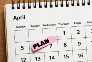 Calendário do mês de abril aberto sobre uma mesa, com um bilhete rosa escrito “PLAN” destacado sobre uma data, representando planejamento antecipado.